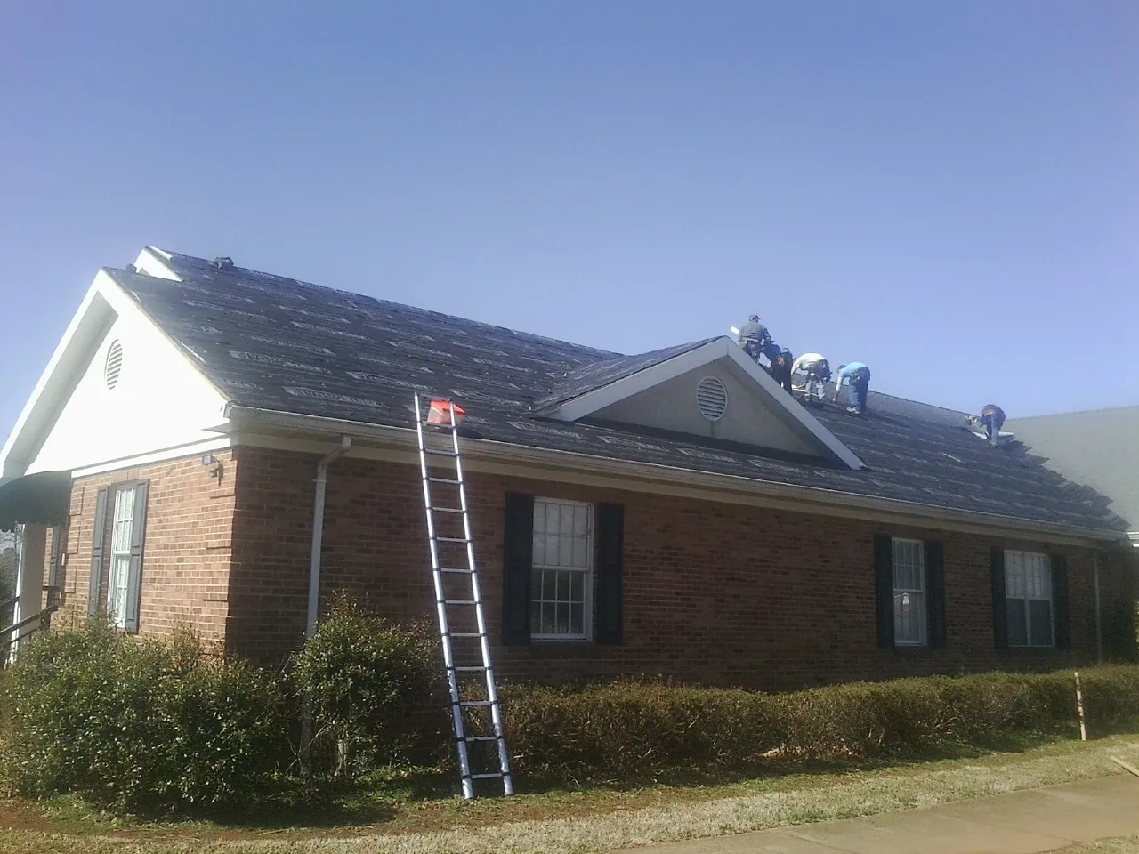 Crew tearing shingles off a brick ranch roof with a ladder set — Emergency Roof Repair work by our crew in Ostrander