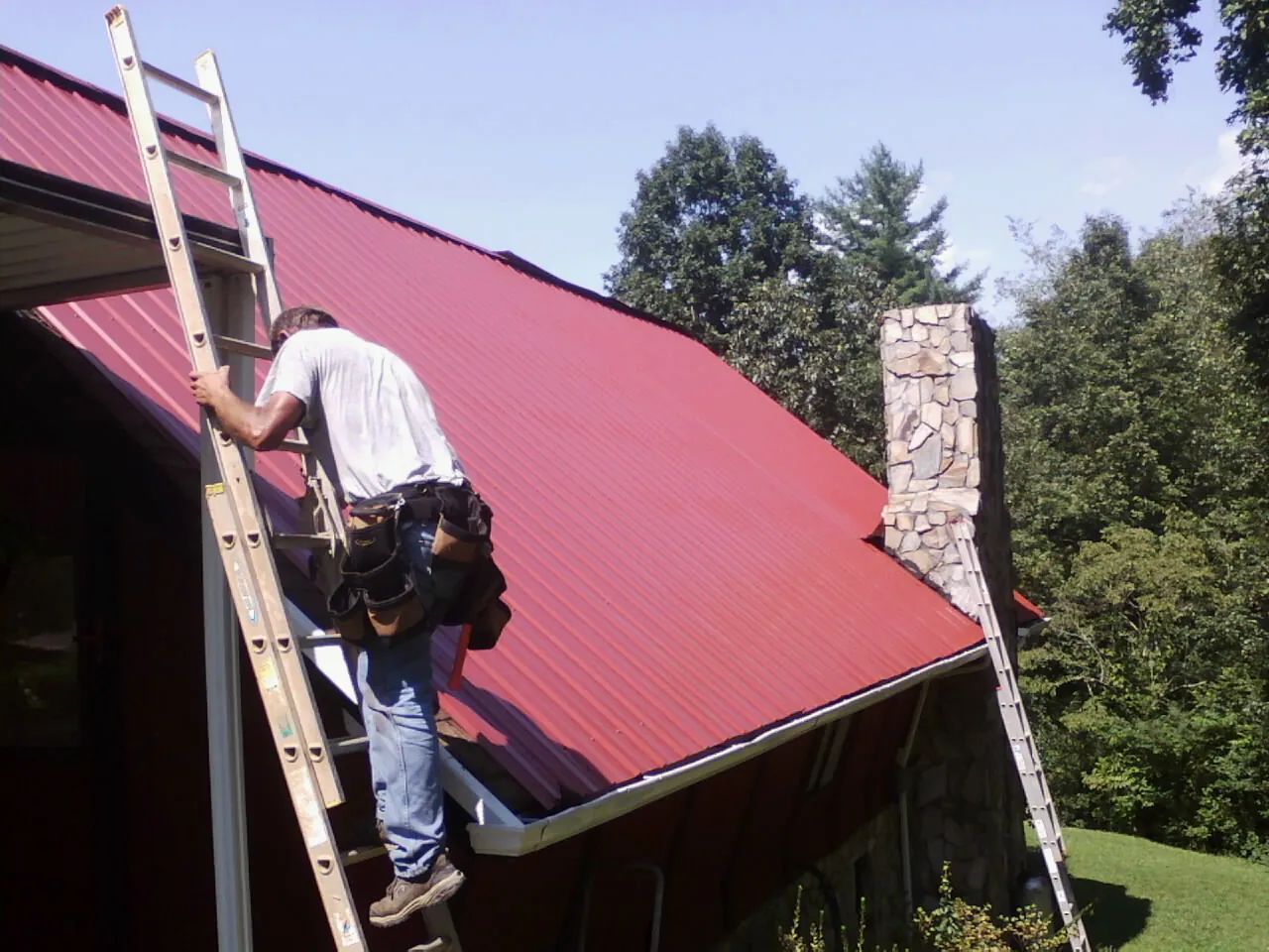 Roofer on a ladder beside a red metal roof and stone chimney — Soffit & Fascia Repair by Roofing Brothers in Ostrander